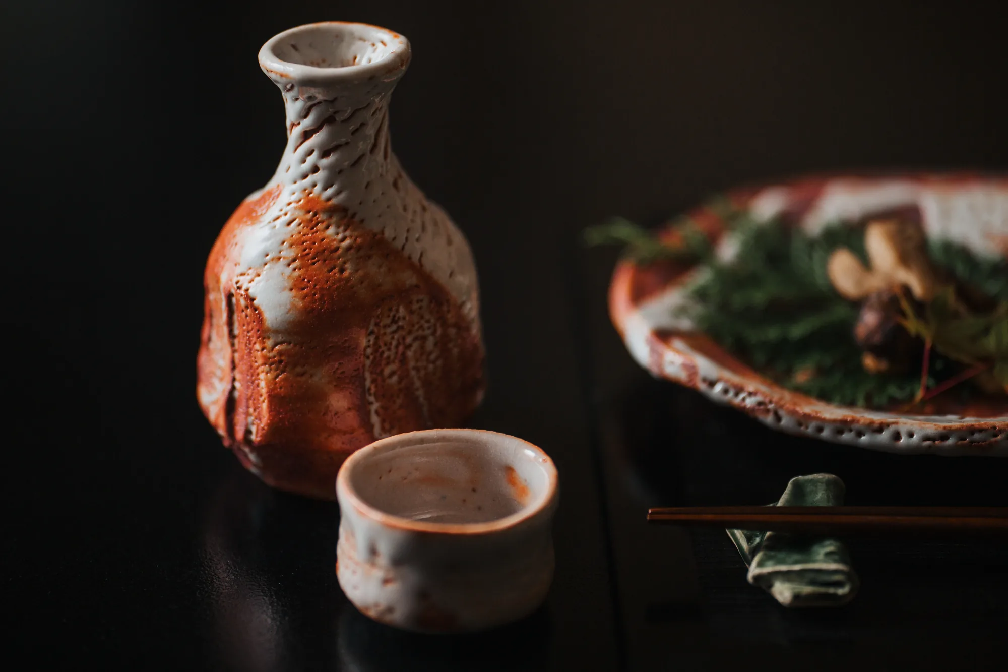 黒い卓に置いた、紅志野の徳利とぐい呑み。奥に料理を盛った紅志野の皿